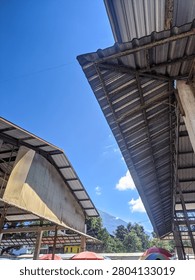 This architectural photograph features a dramatic low-angle perspective of industrial-style corrugated metal roofing. The sharp, parallel lines of the roof panels converge against a vast, deep blue sky, creating a strong sense of height and geometry. The structures appear to be part of a large outdoor market, warehouse, or transit hub shelter. Sunlight illuminates the edges of the metallic surfaces, while the undersides remain in soft shadow. In the distance, the tops of green trees and a hint of a building are visible under the clear atmosphere. This composition highlights the intersection of functional engineering.
