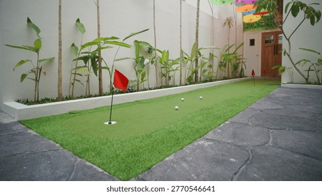 This backyard features a mini golf course with bright green synthetic grass, complete with several holes marked with red flags and golf balls. The ambiance is further enhanced by rows of ornamental plants along the white walls and colorful umbrellas hanging overhead.