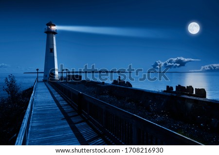 This is a beautiful photo illustration of a dramatic night time scene with a large blue moonrise in a clear sky on a ocean pier with a brightly lit lighthouse beacon and calm ocean waters. 