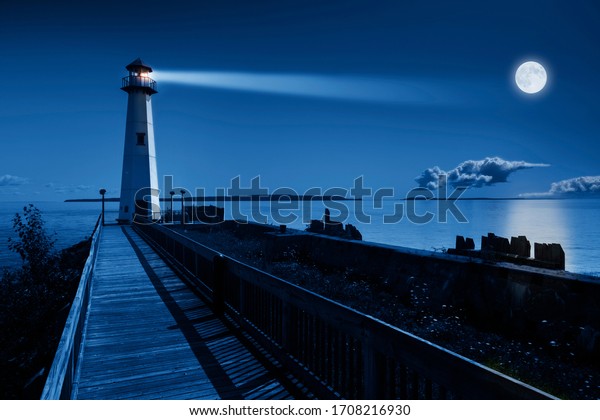 This is a beautiful photo illustration of a dramatic\
night time scene with a large blue moonrise in a clear sky on a\
ocean pier with a brightly lit lighthouse beacon and calm ocean\
waters. 