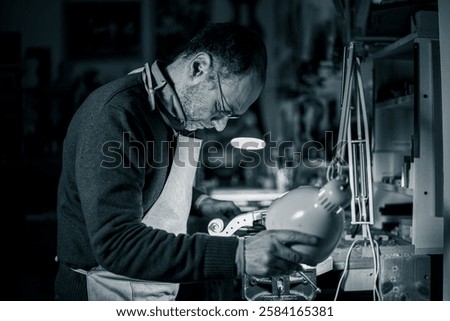 This black and white image captures a craftsman meticulously shaping a violin scroll in his workshop. The scene is bathed in focused light, highlighting his precision and artistry.