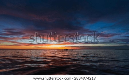 This breathtaking image captures a tranquil ocean scene at sunset, where the sky transforms into a mesmerizing blend of deep blue, purple, orange, and pink hues. The calm water reflects the vivid colors of the sky, creating a harmonious and peaceful atmosphere. At the horizon, a long boat carrying several silhouetted figures glides gracefully across the sea, adding a sense of scale and human connection to the vast natural beauty. The composition perfectly balances the dramatic clouds above with the gentle ripples below, evoking feelings of serenity, adventure, and wonder. This photo is ideal for use in travel, nature, and landscape-themed projects, symbolizing freedom, exploration, and the timeless beauty of the ocean at dusk.