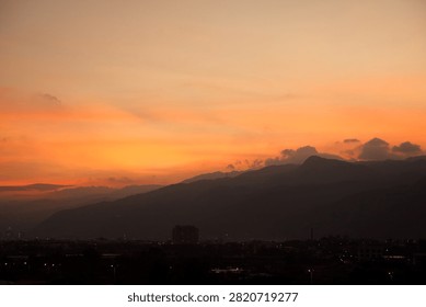 This breathtaking landscape captures a dramatic sunset over a majestic mountain range. The sky glows with vibrant shades of orange, gold, and peach, reflecting the sun's fading warmth. Against this brilliant backdrop, the dark silhouetted peaks and a distant city skyline create a serene, peaceful contrast filled with natural wonder.