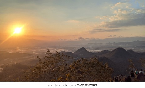 This breathtaking panoramic image captures a radiant sunset casting golden light over a vast mountainous landscape and a distant valley. The rolling hills create depth, while the orange sky and distan