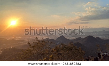 This breathtaking panoramic image captures a radiant sunset casting golden light over a vast mountainous landscape and a distant valley. The rolling hills create depth, while the orange sky and distan