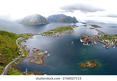 This breathtaking panoramic shot from the summit of Reinebringen offers one of the most famous views in the Lofoten Islands, looking down onto the village of Reine and its surrounding fjords.

The colorful fishing village of Reine is spread across several small islands connected by bridges, appearing as tiny clusters of red and white buildings amidst the vast blue of the Reinefjorden.

Massive, jagged granite mountains rise straight out of the water. The peaks of Olstind and Lofotveggen are partially shrouded in low-hanging clouds, adding a sense of scale and Arctic drama to the landscape of northern Norway.

A winding coastal road (the European E10 scenic Highway) traces the shoreline on the left, highlighting how human settlement clings to the narrow strips of flat land at the base of the cliffs.

From this height, the water transitions from deep navy in the center of the fjord to vibrant turquoise and emerald near the rocky shorelines and submerged reefs.