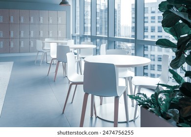 This bright and airy cafeteria area features modern white tables and chairs, providing a clean and minimalist aesthetic. The large floor-to-ceiling windows allow ample natural light to flood the space - Powered by Shutterstock