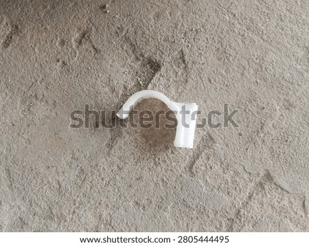 This is a broken white plastic clip, specifically a PVC pipe clamp or cable saddle, designed to secure pipes or cables to walls or floors. Only the curved upper section and one mounting lug remain; the other side is snapped off, leaving rough, uneven edges. The surface is slightly soiled with dust and grit, lying on a rough, grey concrete floor marked by fine cracks and stains.