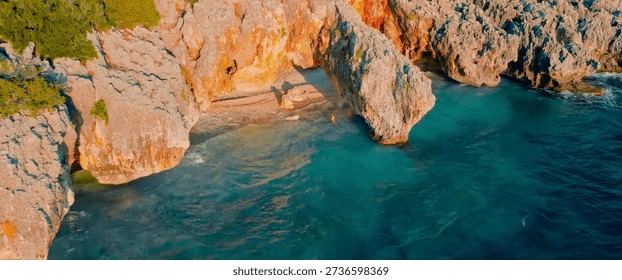 This captivating aerial perspective features a dramatic natural rock arch overhanging a secluded, pebble-strewn cove where shimmering emerald and deep blue waters meet the rugged limestone cliffs. Bathed in the warm, golden glow of the late afternoon sun, the scene highlights the sharp textures of the rocky terrain and the lush greenery clinging to the precipice, while a solitary figure stands near the arch, emphasizing the vast, tranquil scale of this hidden Mediterranean gem.