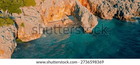 This captivating aerial perspective features a dramatic natural rock arch overhanging a secluded, pebble-strewn cove where shimmering emerald and deep blue waters meet the rugged limestone cliffs. Bathed in the warm, golden glow of the late afternoon sun, the scene highlights the sharp textures of the rocky terrain and the lush greenery clinging to the precipice, while a solitary figure stands near the arch, emphasizing the vast, tranquil scale of this hidden Mediterranean gem.