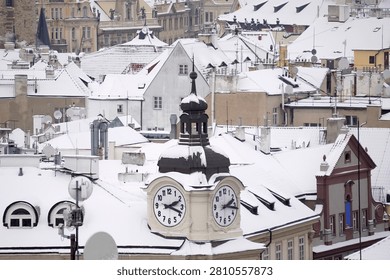 This captivating high angle winter photograph focuses on a beautiful vintage clock tower rising from the center of a historic European city. The elegant architectural tower features two large white clock faces showing the time, topped with a distinctive dark baroque dome capped by a light dusting of fresh winter snow. In the background, a dense maze of traditional town rooftops, residential facades, dormer windows, chimneys creates a classic urban tapestry. Every surface, roof tile, architectural ledge is thickly covered in pristine white snow, capturing a peaceful, quiet atmosphere during the cold season. The architectural style represents centuries of European history, combining classical gables with ancient town layout elements. The color palette remains soft and elegant, dominated by the bright white of the winter snow, the pale beige and pink hues of the historical building facades, the dark metallic tones of the tower roof structure. Soft natural daylight illuminates the scene uniformly, emphasizing the fine details of the clock mechanism dials, the textures of the snowy roofs, the cozy arrangement of old town houses. 