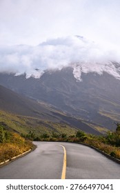 This captivating image captures a winding road cutting through a verdant landscape, leading towards a majestic mountain partially shrouded in clouds. The road, prominently featured, is smooth and reflects the ambient light, emphasizing its path into the distance. A striking yellow line marks the center, guiding the eye towards the imposing mountain. The surrounding terrain is lush with vegetation, adding a vibrant contrast to the gray tones of the mountain. The mountain itself, a blend of rocky textures and patches of snow, looms large, its peak obscured by a thick layer of clouds, lending an air of mystery and grandeur. The image evokes a sense of adventure and exploration, suggesting a journey through untouched natural beauty. The lighting is soft and diffused, creating a tranquil and atmospheric mood. This scene is located in Chile, where nature’s beauty meets the adventurous spirit.