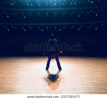 This captivating image portrays a lone performer standing center stage in a grand, dimly lit theater. The dancer faces an expansive, empty auditorium illuminated by soft blue and white stage lights, w