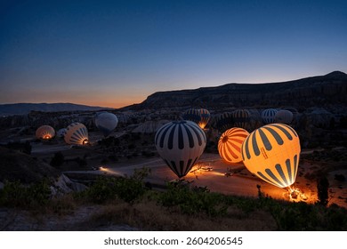 This captivating image showcases a tranquil early morning in Cappadocia, Turkey, as vibrant hot air balloons prepare for their ascent. Illuminated from within, the balloons emit a warm glow against th