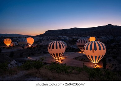 This captivating image showcases a tranquil early morning in Cappadocia, Turkey, as vibrant hot air balloons prepare for their ascent. Illuminated from within, the balloons emit a warm glow against th