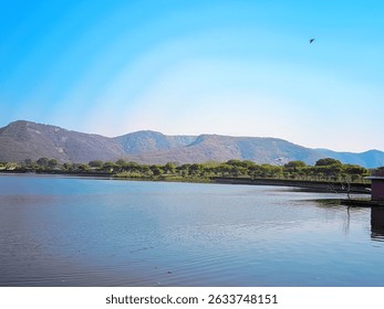 This captivating image showcases the tranquil beauty of Man Sagar Lake in Jaipur, Rajasthan, India, framed by the majestic Aravalli Hills under a clear blue sky. The serene waters of the lake reflect 