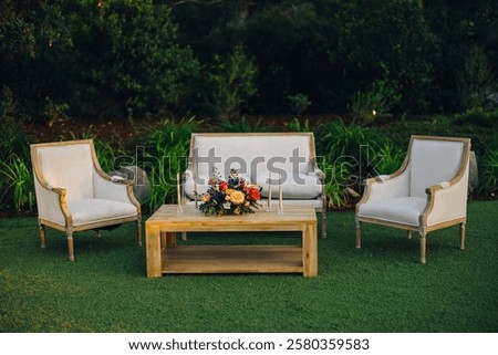This charming outdoor area features a stylish sofa, comfy armchairs, and a floral centerpiece on a wooden table, all surrounded by lush greenery, creating a serene space for relaxation and gatherings