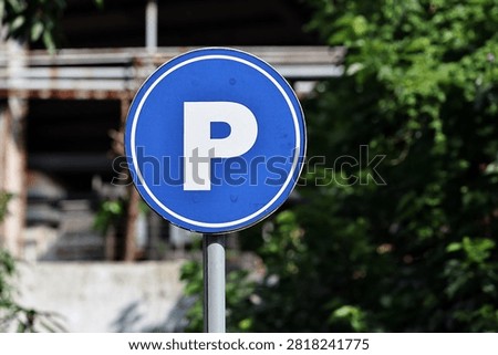 This circular traffic sign indicates a designated parking area, featuring a bold white letter P on a solid blue background. Mounted on a metallic pole, the sign stands outdoors surrounded by lush green foliage and an urban building structure. It serves as a clear visual guide for drivers, ensuring organized vehicle parking and smooth traffic flow.