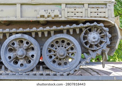 This close-up captures the rugged lower section of a military armored vehicle featuring worn steel tracks and heavy-duty road wheels. The frame exhibits an olive-drab finish, showing signs of weathering, light rust, and historical service. Two large, circular driving wheels dominate the mid-ground, characterized by a complex pattern of ventilation holes and central hubs. To the right, a smaller, intricate sprocket gear engages directly with the metal track links, demonstrating the mechanical complexity of the propulsion system. The massive tread consists of thick, connected plates designed for traversing difficult terrain, while the upper structure provides a sturdy platform for the tank's chassis. Natural sunlight illuminates the textured surfaces, highlighting the patina of the aged metal and the shadows cast within the deep recesses of the undercarriage. Green foliage is visible in the background, suggesting the equipment is currently displayed in an outdoor exhibition or park setting. The composition emphasizes the industrial power and engineering durability inherent in heavy combat machinery. Every bolt, link, and rim reflects a history of operation, with the stark contrast between the dull, earthy tones of the hardware and the vibrant green vegetation creating a compelling visual narrative. This mechanical assembly serves as a testament to battlefield resilience and mid-twentieth-century design principles, offering a detailed perspective on the structural components that once allowed such immense machines to dominate the landscape during intensive operations.