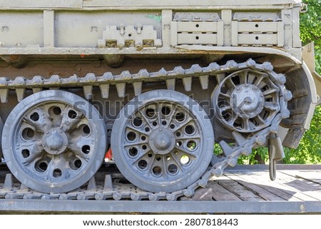 This close-up captures the rugged lower section of a military armored vehicle featuring worn steel tracks and heavy-duty road wheels. The frame exhibits an olive-drab finish, showing signs of weathering, light rust, and historical service. Two large, circular driving wheels dominate the mid-ground, characterized by a complex pattern of ventilation holes and central hubs. To the right, a smaller, intricate sprocket gear engages directly with the metal track links, demonstrating the mechanical complexity of the propulsion system. The massive tread consists of thick, connected plates designed for traversing difficult terrain, while the upper structure provides a sturdy platform for the tank's chassis. Natural sunlight illuminates the textured surfaces, highlighting the patina of the aged metal and the shadows cast within the deep recesses of the undercarriage. Green foliage is visible in the background, suggesting the equipment is currently displayed in an outdoor exhibition or park setting. The composition emphasizes the industrial power and engineering durability inherent in heavy combat machinery. Every bolt, link, and rim reflects a history of operation, with the stark contrast between the dull, earthy tones of the hardware and the vibrant green vegetation creating a compelling visual narrative. This mechanical assembly serves as a testament to battlefield resilience and mid-twentieth-century design principles, offering a detailed perspective on the structural components that once allowed such immense machines to dominate the landscape during intensive operations.