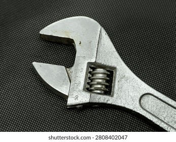 This is a closeup, highangle shot of an adjustable wrench. The tool is made of metal and has a textured handle. The jaws of the wrench are open, revealing the adjustable mechanism and measurement markings. The background is a dark, woven fabric providing a contrasting texture to the smooth metal of the tool.