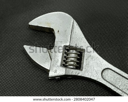 This is a closeup, highangle shot of an adjustable wrench. The tool is made of metal and has a textured handle. The jaws of the wrench are open, revealing the adjustable mechanism and measurement markings. The background is a dark, woven fabric providing a contrasting texture to the smooth metal of the tool.