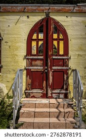 This close-up image captures the striking red double doors of the weathered yellow church on Jost Van Dyke, featuring peeling paint that reveals the wood underneath, arched tops with multi-pane glass windows.