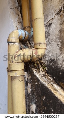 This close-up photograph captures a section of old, weathered plumbing pipes mounted on a deteriorating wall. The yellow pipes, once coated in smooth paint, now show heavy signs of peeling, rust, and 