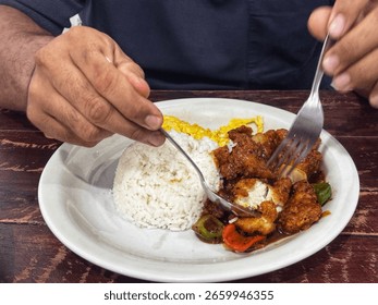 This close-up shot captures a person's hands using a fork and spoon to enjoy a meal served on a white plate. The plate holds a generous portion of white rice, an omelet (or fried egg) that's partially