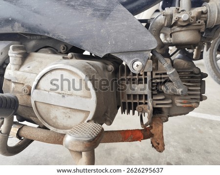 This is a close-up shot of a motorcycle engine. The engine block and components appear worn and dusty. A rusty exhaust manifold and other mechanical parts are visible.
