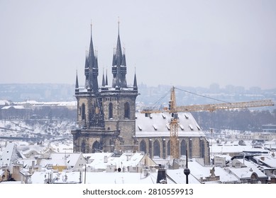 This compelling high angle winter cityscape captures the iconic dark gothic spires of the historic Church of Our Lady before Tyn rising majestically over the snow covered rooftops of Prague. The monumental church architecture features two prominent twin towers decorated with intricate stone pinnacles, arched windows, medieval details, all lightly dusted with fresh winter snow. In the foreground, a dense network of traditional town houses, residential roofs, small chimneys extends across the landscape, completely blanketed in a thick sheet of pristine white seasonal frost. A prominent yellow construction crane stands next to the church structure, representing urban development, reconstruction, restoration processes within the ancient heritage site. The background reveals the snow covered hills, bare winter trees, distant misty urban outlines under an overcast grey sky that diffuses natural light evenly across the entire frame. The color scheme is cold and beautifully minimalist, dominated by the bright white tones of the fresh snow, the dark weathered stone of the gothic towers, the soft neutral greys of the winter atmosphere, the contrasting bright orange hue of the crane. 