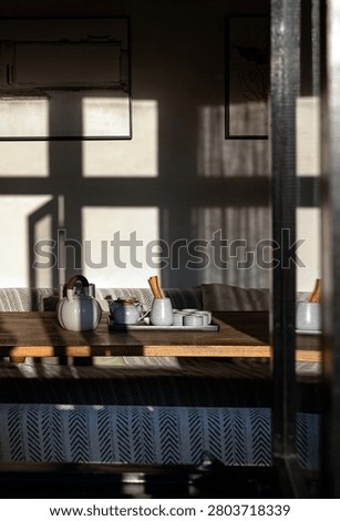 This composition captures a beautiful interplay of light and shadow, filtering through a window frame to create dramatic, grid-like patterns on a neutral wall. In the foreground, a finely crafted ceramic teapot and an array of delicate teacups rest on a wooden tabletop, creating an inviting scene of traditional, understated elegance.