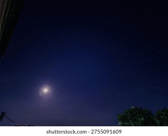 In this composition, the celestial meets the terrestrial. The focal point is a luminous full moon hanging low in the sky, surrounded by a soft, purple-hued glow that bleeds into the dark indigo of the night. Unlike the first image, this scene provides a sense of place. The silhouettes of lush green treetops in the bottom right and the corner of a roof in the top left frame the sky, grounding the viewer in a specific moment on Earth.
​The stars here are more sparse but appear sharp against the darker backdrop, creating a balanced contrast with the dominant light of the moon. The overall atmosphere is one of peaceful solitude—the kind of view one might find while stepping outside on a quiet evening. It captures that perfect, fleeting balance where the brilliance of the moon is strong enough to light the landscape while still leaving room for the stars to shine.