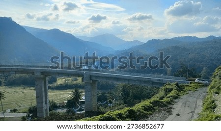 This concrete flyover cuts through hills with sturdy support pillars, serving as a route to bypass steep inclines. The gray concrete structure gives an industrial, modern look, contrasting with the surrounding greenery. Access roads flank the bridge on the hillside. The background features layered hills with thin fog, a lush valley, and residential homes at the valley base. The atmosphere is calm with natural lighting casting shadows on the bridge pillars.