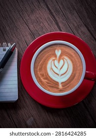This cozy flat-lay image features a warm cup of coffee served in a bright red ceramic mug and matching saucer, resting on a rustic wooden table. The drink is beautifully adorned with intricate heart-shaped latte art. Beside the cup lies a small, spiral-bound lined notepad with a sleek black pen placed on top, evoking a quiet, productive atmosphere perfect for journaling, planning, or enjoying a mindful break.
