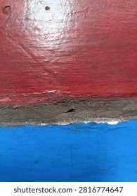 This detailed, close-up composition, captured in natural daylight, highlights the tactile textures and imperfections of aged, outdoor wood. The top horizontal section consists of a heavily-used, faded crimson plank with significant paint wear, chipping, and scratches revealing the rough wood grain beneath. This red layer is heavily distressed, showing signs of prolonged exposure to sun, rain, and time. Below the crimson section lies a rugged, unpainted strip of dark grey wood, rough, splintered, and deeply cracked, acting as a natural, aged division. This grey wood features a prominent vertical fissure and rough, pitted surface, suggesting heavy use on a traditional boat or old building. The bottom horizontal section displays a contrasting, yet equally weathered, cobalt blue plank. The blue paint is also peeling, cracked, and scratched, but retains a more vivid hue compared to the red, with a visible texture where previous layers might have been sanded. The two colored planks meet the central grey piece unevenly, creating a dynamic, jagged boundary. The depth of field is shallow, focusing sharply on the textures and imperfections along the junction and the grain. The photograph's style is unrefined and gritty, capturing authentic wear.