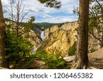 This is a distant view of Yellowstone Falls, located in Yellowstone National Park in Wyoming. Steep tiered rock cliffs encompass the falls, and mountains are seen way in the distance.