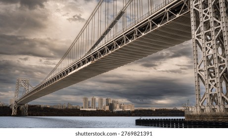 This dramatic photograph captures a massive suspension bridge spanning a wide body of water. The imposing steel structure cuts across the frame, contrasting sharply against the moody, overcast sky and the distant urban skyline. It evokes themes of engineering prowess, travel, connection, and industr