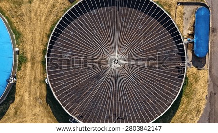 This dramatic top-down aerial photograph provides an extraordinary close-up view of a large open circular biogas digester tank, showcasing its impressive radial spoke structure and dark organic contents. The geometric pattern creates a powerful visual impact, with adjacent bright blue dome digesters and surrounding infrastructure visible for context. The high-angle perspective reveals technical details and the scale of industrial bioenergy equipment.