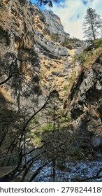 This dramatic vertical view captures a towering, steep canyon wall towering over a rocky riverbed. The immense geological formation features prominent textured rock layers with warm shades of orange, tan, and dark grey. Various trees and wild vegetation cling tightly along the narrow crevices of the cliff face, while a blue sky with soft white clouds peeks out at the very top. In the foreground, dense, dark branches frame a small wooden footbridge on the left, emphasizing the grand scale and natural majesty of this remote valley.
