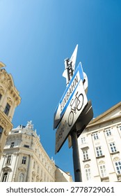 This dynamic low-angle photograph captures a cluster of traffic signs mounted on a grey pole under a vibrant, cloudless blue sky. The prominent sign is a blue rectangular 