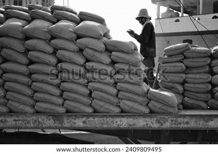 In this dynamic scene at Sunda Kelapa Port Jakarta Indonesia, a worker praying first while working to loads sacks onto trucks against the backdrop of a bustling maritime environment