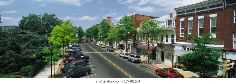 This Is The Eastern Shore Of Maryland. It Typifies Small Town America Or Main Street USA. We See Shop Fronts On A Tree Lined Street. Cars  Are Parked In Front Of Shops On Either Side Of The Street.