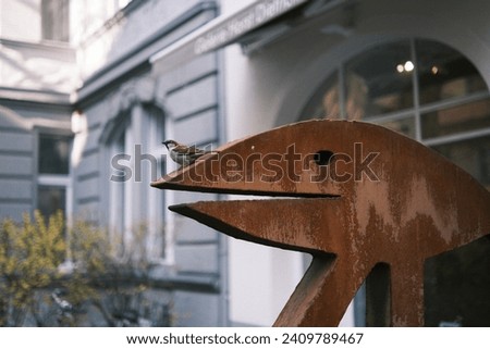 This engaging photograph showcases a sparrow resting on an abstract metal sculpture, set against the backdrop of a German city's streetscape.