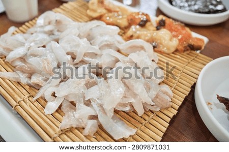 This enticing culinary visual presents a generous serving of fresh filefish sashimi elegantly arranged on a traditional bamboo mat The translucent white slices of raw fish highlight their premium quality and firm chewy texture making them a highly sought after delicacy in South Korean dining In the background a glimpse of vibrant orange sea squirt adds to the authentic seafood experience This popular dish is famously paired with local spirits like soju for an authentic nightlife vibe Ideal for Asian restaurant menus gastronomy features and commercial projects promoting fresh marine cuisine