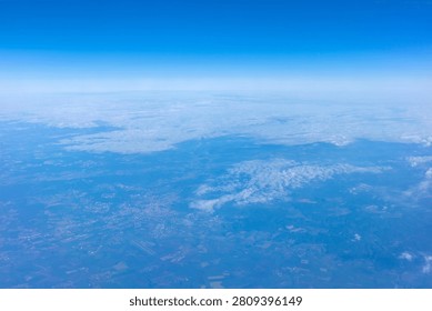 This expansive aerial perspective captures a serene, sunlit landscape blanketed beneath a vast, luminous blue sky dotted with gentle clouds. From high above, the terrain unfolds in a patchwork of soft textures, where subtle variations in color indicate rolling hills, dense patches of woodland, and scattered human settlements. Small urban areas appear as clusters of muted tones, intersected by thin, linear features that suggest roadways or perhaps infrastructure. A prominent runway is clearly visible, cutting through the terrain, highlighting the human presence amidst the natural expanse. Wispy, white cumulus formations drift lazily across the horizon, casting faint, ethereal shadows that dance upon the ground below, lending a sense of immense depth and calm to the entire scene. The transition from the clear, brilliant atmosphere near the zenith to the slightly hazy, distant horizon creates a powerful feeling of height and tranquility. Sunlight bathes the entire view in a cool, atmospheric glow, emphasizing the peaceful isolation of flying through the upper air. It is a moment of quiet observation, looking down upon the world as it stretches out in endless, intricate detail under a boundless dome of cerulean light.