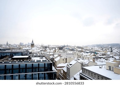 This extensive high angle panoramic winter photograph captures a wide urban landscape of a European city blanketed under fresh white snow. The modern dark glass building facade in the lower left corner creates a stark contemporary contrast against the vast sea of traditional historical rooftops stretching toward the horizon. Numerous ancient tower spires, church domes, historical architectural details emerge from the dense residential grid, illustrating the rich cultural heritage of this capital city. A thick uniform layer of pristine winter snow covers every visible roof, terrace, street line, building structure below. The sky occupies a significant portion of the frame, filled with dense overcast grey and white winter clouds that create a soft diffused natural illumination over the entire landscape. In the far distance, snow covered rolling hills and bare trees mark the boundary of the urban area under the hazy winter horizon. 