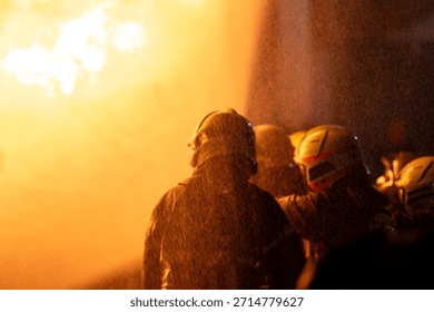 This is an extremely close-up and dynamic shot capturing the intense action of firefighting. A wall of bright, turbulent orange and yellow flames dominates the left side of the frame, indicating a large fire. On the right, partially obscured and visible primarily by their protective gear, are several firefighters. The most prominent figure is wearing a light-colored (possibly yellow or white) helmet with a clear face shield. Another figure next to them has a red face shield, intensely illuminated by the blaze. The entire scene is enveloped in a thick, warm, orange glow and filled with countless water droplets or ash particles suspended in the air, emphasizing the extreme heat and proximity to the fire.