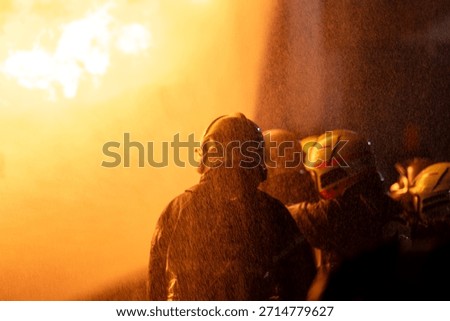 This is an extremely close-up and dynamic shot capturing the intense action of firefighting. A wall of bright, turbulent orange and yellow flames dominates the left side of the frame, indicating a large fire. On the right, partially obscured and visible primarily by their protective gear, are several firefighters. The most prominent figure is wearing a light-colored (possibly yellow or white) helmet with a clear face shield. Another figure next to them has a red face shield, intensely illuminated by the blaze. The entire scene is enveloped in a thick, warm, orange glow and filled with countless water droplets or ash particles suspended in the air, emphasizing the extreme heat and proximity to the fire.