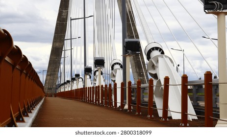 This eye-level perspective of a modern cable-stayed bridge emphasizes urban development, connectivity, and structural integrity. The empty pedestrian pathway, framed by a distinct orange railing and intricate white support cables, symbolizes a journey or path towards the future in a metropolitan setting. Ideal for showcasing engineering marvels, infrastructure projects, or urban planning in corporate business, travel, and public works contexts